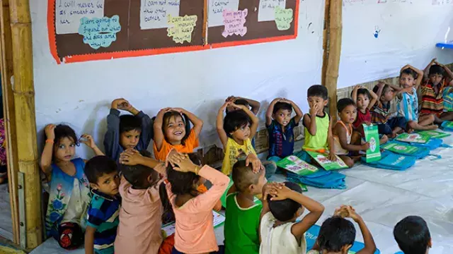 A group of children sitting on the floor in a classroom, with some raising their hands.